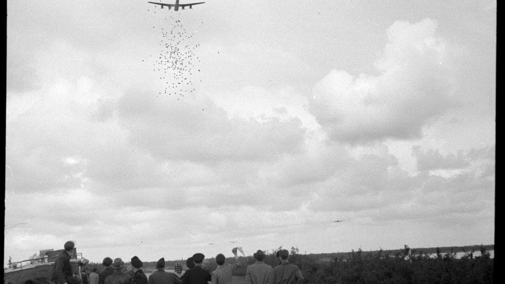 zwart-witfoto van een bommenwerper die voedselpakketten dropt terwijl mensen toekijken.