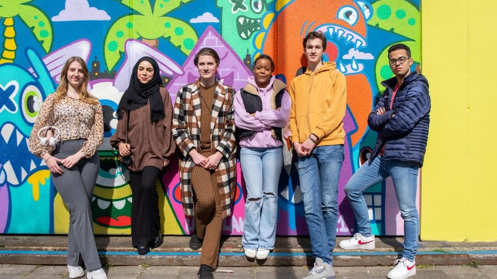 Groep van 6 jongeren leunt zelfverzekerd tegen een muur met graffiti.