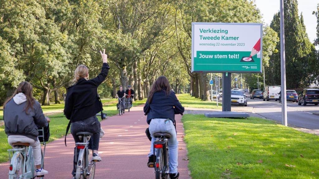 Drie fietsers fietsen bij een bord waarop wordt opgeroepen om te stemmen. Foto: Rhalda Jansen.